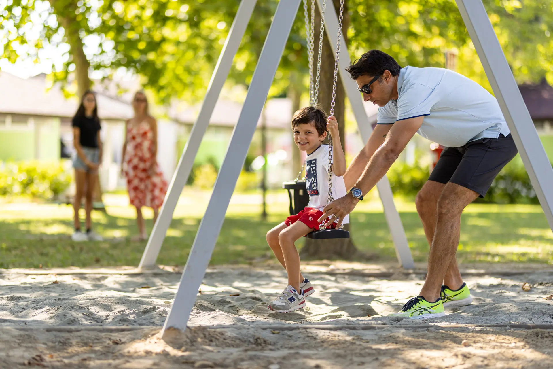 Bambino sorridente su un’altalena mentre un adulto lo spinge nel parco giochi sabbioso e ombreggiato del Villaggio San Francesco.