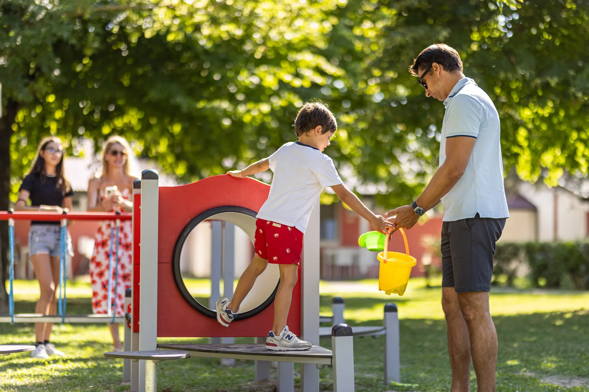 Bambino su un percorso giochi che riceve un secchiello da un adulto nel parco ombreggiato del Villaggio San Francesco.