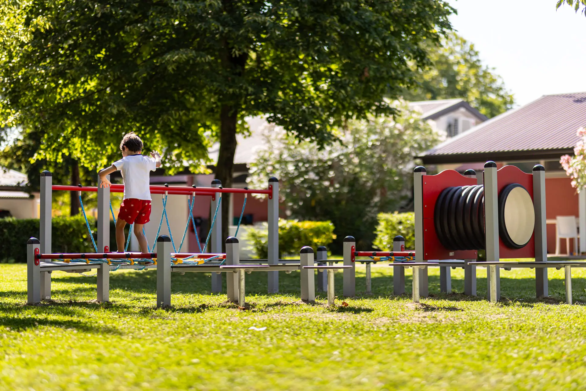 Bambino che percorre un piccolo percorso a ostacoli nel parco giochi del Villaggio San Francesco, prato verde e alberi in ombra.