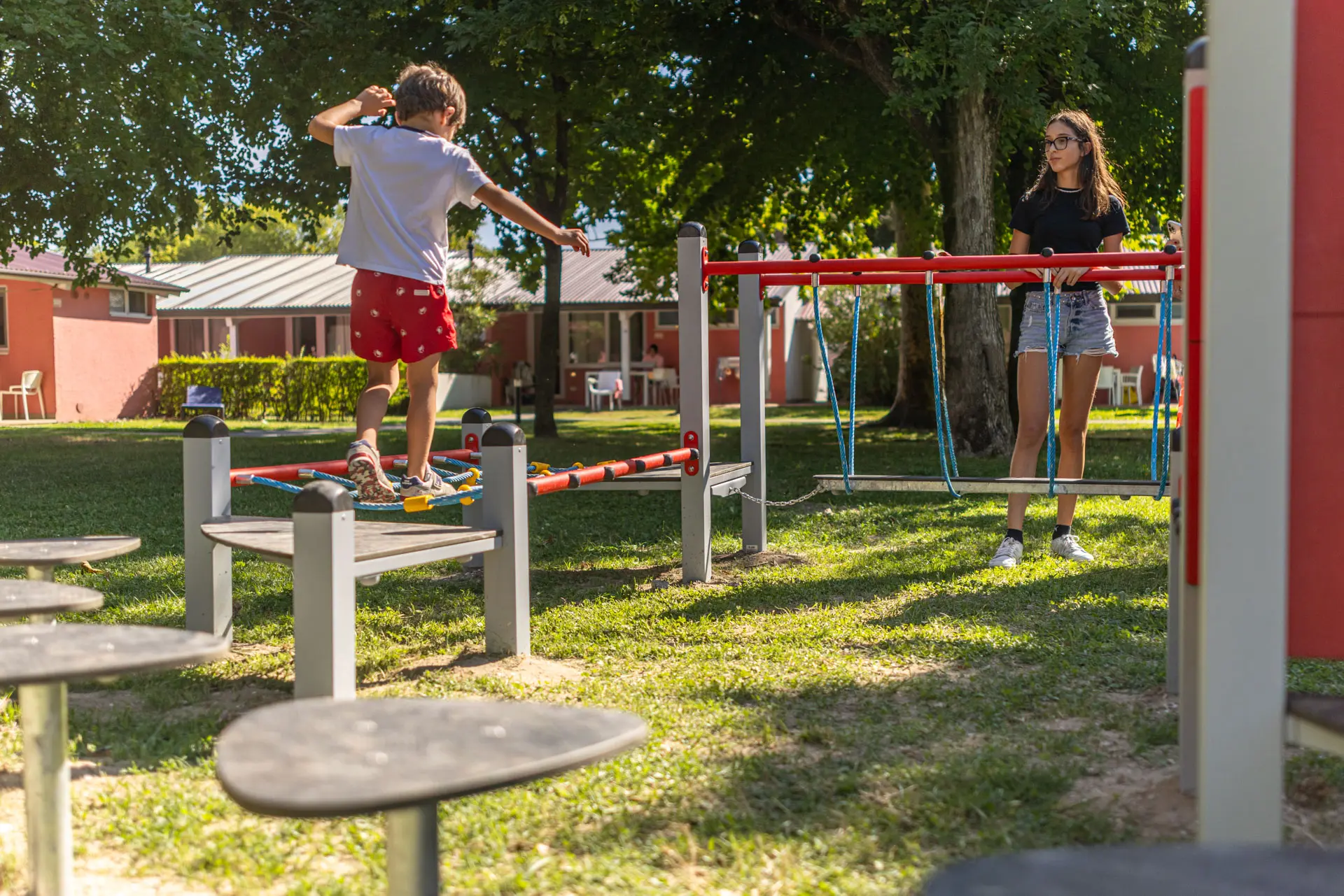 Bambino che attraversa un percorso a ostacoli sospeso nel parco giochi ombreggiato del Villaggio San Francesco, con una ragazza sullo sfondo.